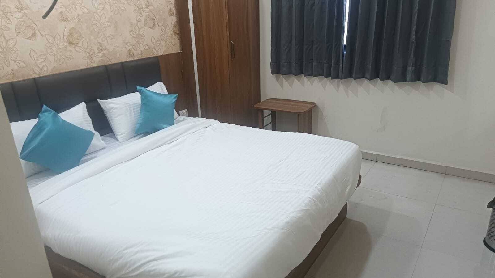 Una cama bien hecha con ropa blanca y almohadas azules, un cabecero de madera, un taburete pequeño de madera y una ventana con cortinas oscuras.