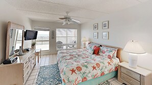Ein Schlafzimmer mit einem Bett, einem Deckenventilator, einem Fernseher, einer Kommode, einer Lampe und einem Fenster mit Jalousien.