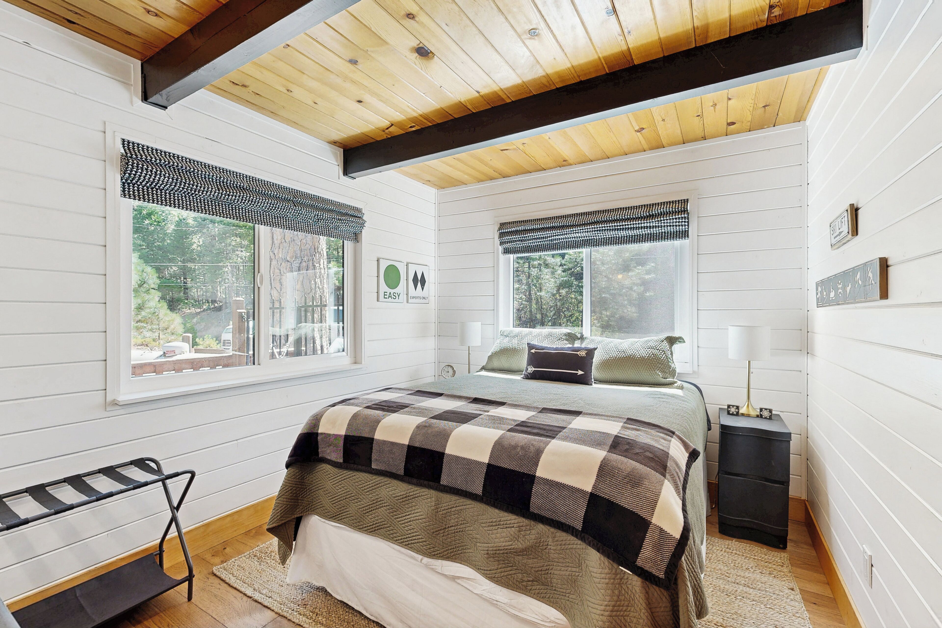 Une chambre à coucher avec un lit, deux fenêtres avec des rideaux, une table de chevet avec une lampe et un banc près de la fenêtre.