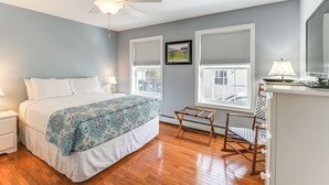 Ein Schlafzimmer mit einem Bett, einem Deckenventilator, einem Fenster mit Jalousien, einem Sessel und einer Kommode.