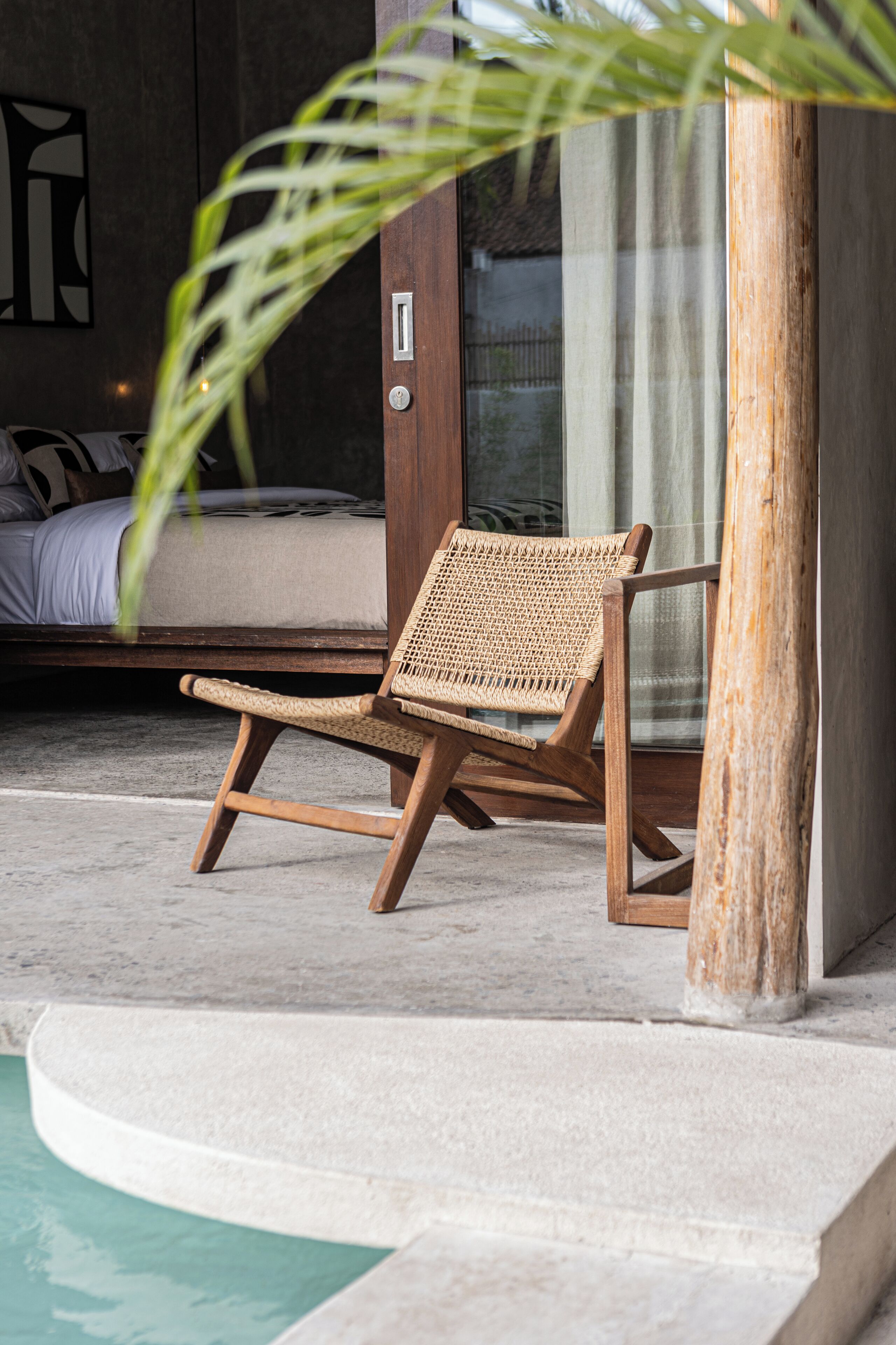 A wooden lounge chair by a poolside, with a concrete floor and a door in the background.