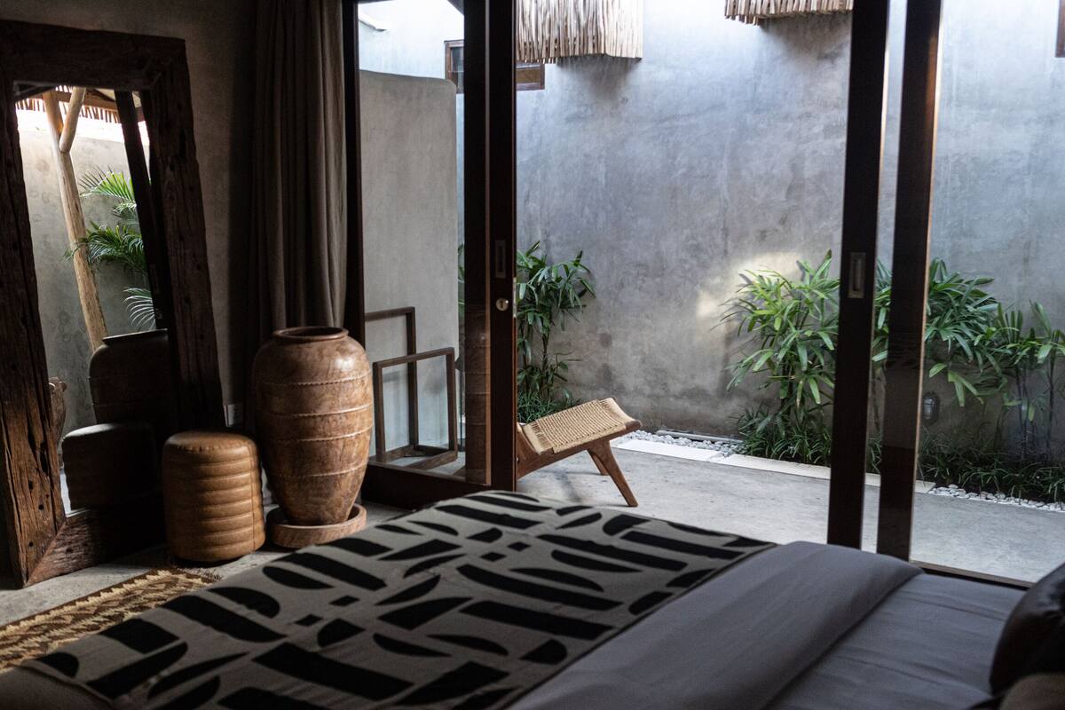 A bedroom with a bed, a wooden chair, a large vase, and a view of a garden through a sliding door.