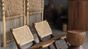 A room with two wooden lounge chairs, a large woven wall hanging, a tall vase with a red flower, and a tall wooden sculpture. - Kano Twin (Canggu)