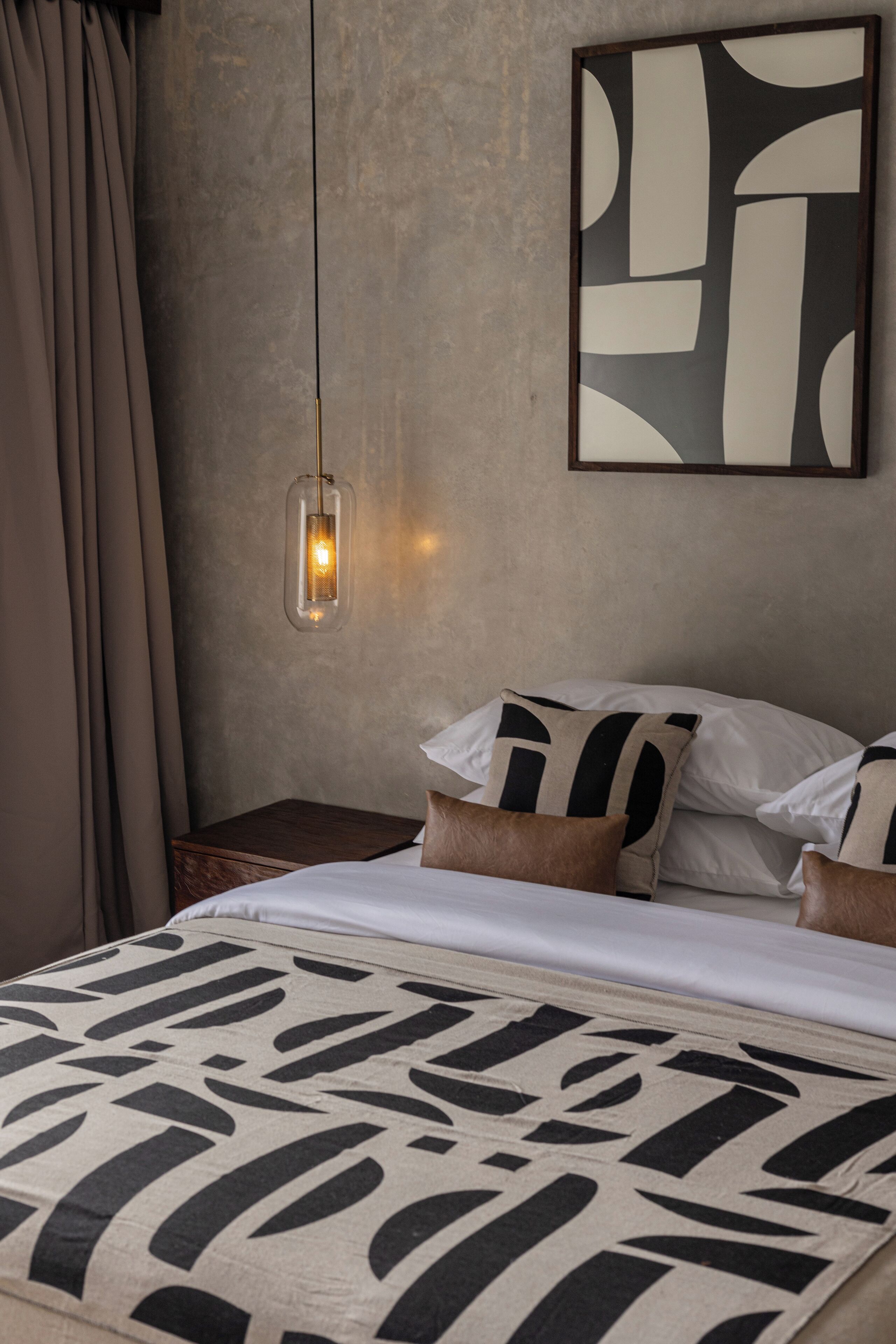 A bedroom with a bed, pillows, a nightstand, a wall-mounted light, and a geometric artwork.