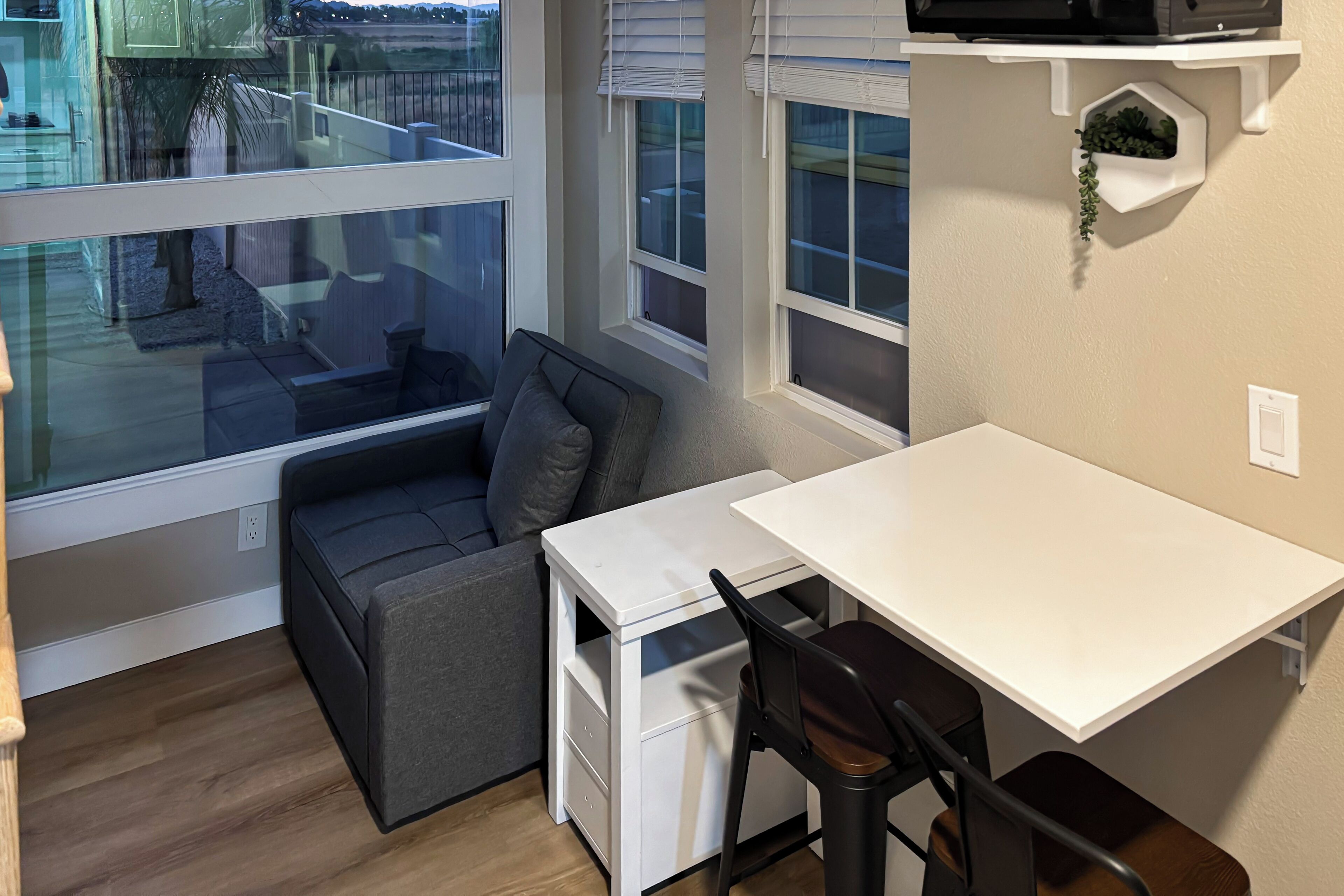 Une pièce de taille modeste, avec un bureau blanc, un canapé gris et une fenêtre donnant sur un balcon.