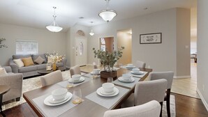 Un coin repas avec une longue table dressée pour six personnes, entourée de six chaises. On aperçoit un canapé et une table basse au fond.
