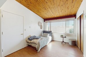 A bedroom with a wooden ceiling, a bed with a headboard, a window with blinds, a small table with a plant, and a trash can. - Poolside Oasis! Phoenix Group Sanctuary w/ Patio (Phoenix)