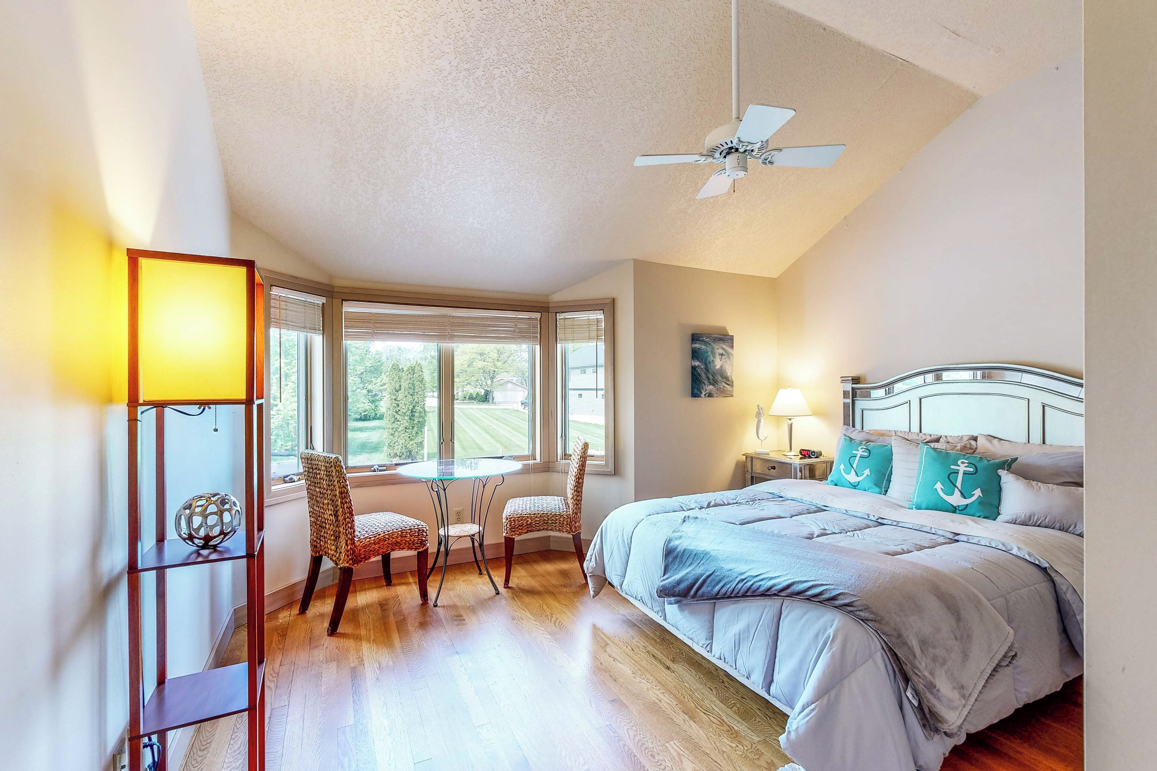 Une chambre à coucher comprenant un lit, un bureau, deux chaises, un ventilateur de plafond et une fenêtre avec des stores.