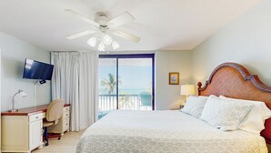 Camera d'albergo con un letto, una scrivania con una sedia, una TV, una ventola a soffitto, un balcone con vista sull'oceano e un comodino con una lampada.