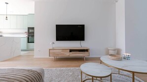 A modern living room with a flat-screen TV mounted on a white wall, a wooden TV stand with a lamp, a round marble coffee table, and a beige armchair. - Brand new house Sable d'Or  - Oostduinkerke (Koksijde)