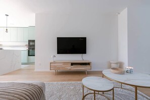 Una sala de estar moderna con un televisor de pantalla plana montado en una pared blanca, un mueble de televisión de madera con una lámpara, una mesa de centro redonda de mármol y un sillón color beige.