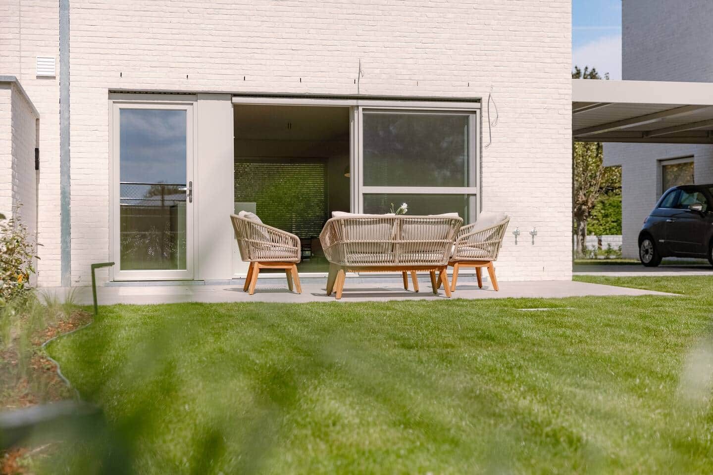 Ein modernes Haus mit weißer Fassade und einer Schiebetür zu einer Terrasse mit Korbmöbeln.