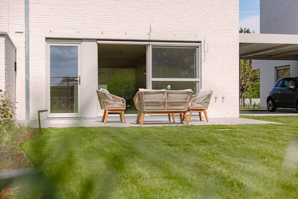 Ein modernes Haus mit weißer Fassade und einer Schiebetür zu einer Terrasse mit Korbmöbeln.