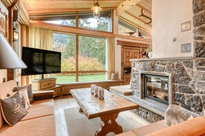 A cozy living room with a stone fireplace, a large window with a view of trees, a flat-screen TV, and a wooden table. - Luxurious and Calm Central Hideaway (Tschagguns)