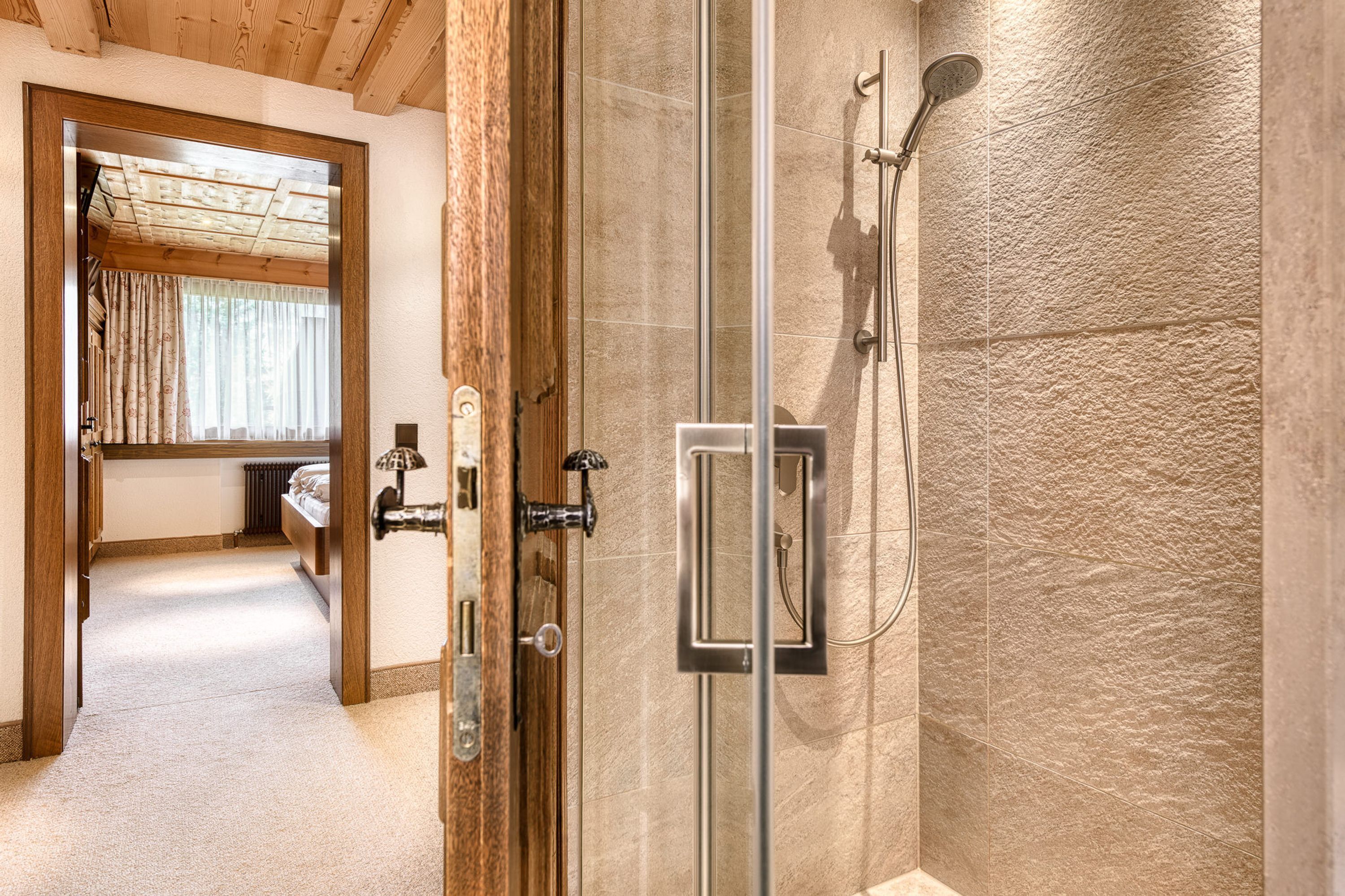 Hotelkamer met houten plafond, een douchecabine van glas en uitzicht op een slaapkamer met bed en gordijnen.