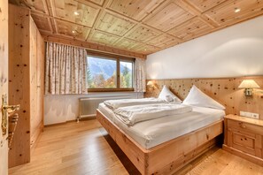 Quarto com painéis de madeira, uma cama grande, janela com cortinas, radiador e mesa de cabeceira com abajur.