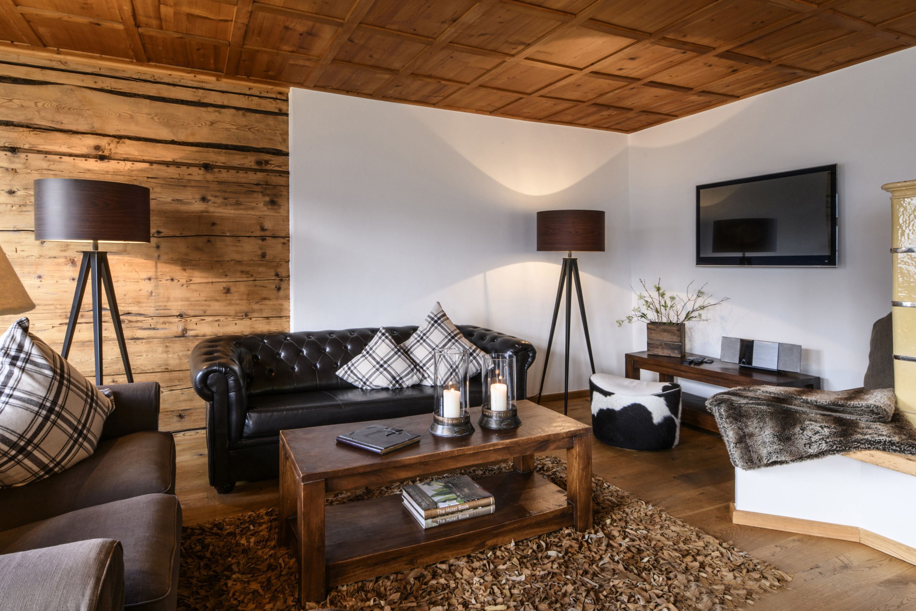 Ein Wohnzimmer mit Holzboden, einem schwarzen Ledersofa, einem Couchtisch mit Büchern, einer Stehlampe und einem an der Wand befestigten Fernseher.