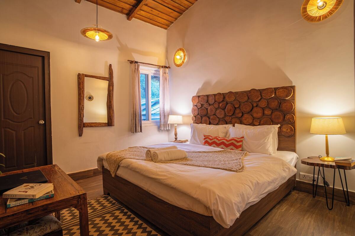 Une chambre à coucher avec une tête de lit en bois, un lit recouvert de draps blancs, une table de chevet avec une lampe, une fenêtre avec des rideaux et une porte en bois.