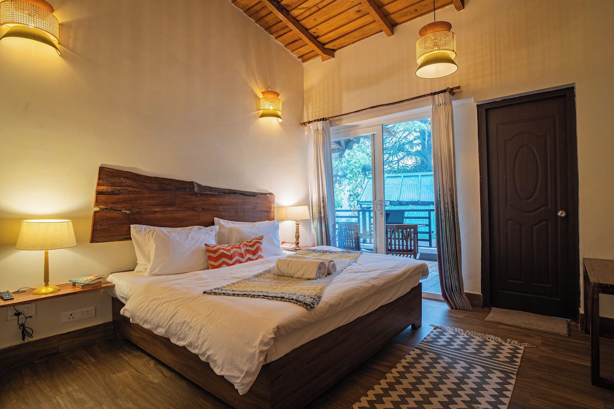 Une chambre à coucher avec une tête de lit en bois, un lit recouvert de draps blancs, deux lampes de chevet et une vue sur l’extérieur par une porte.
