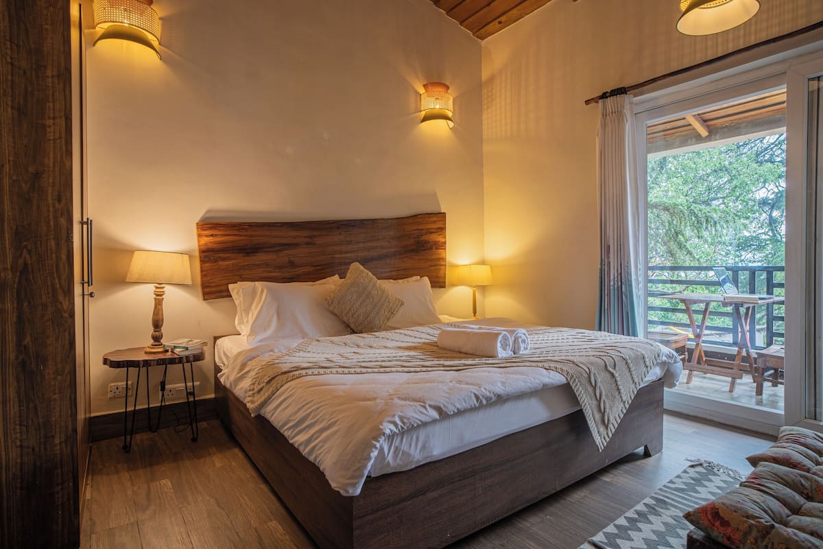 Une chambre à coucher avec une tête de lit en bois, deux lampes de chevet, un lit avec du linge de lit blanc, une petite table avec une lampe et une vue sur une terrasse extérieure.