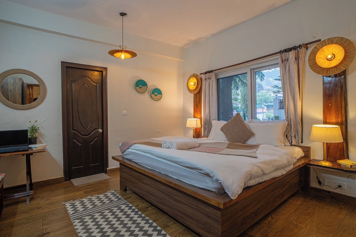 Une chambre à coucher avec un lit en bois, un bureau avec un ordinateur portable, une fenêtre avec des rideaux et une lampe suspendue.