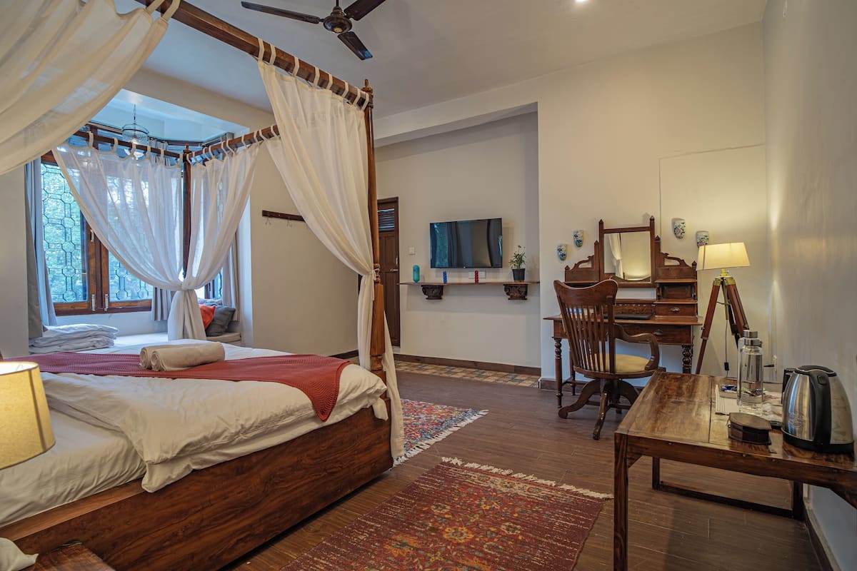 Une chambre à coucher avec un lit à baldaquin, un bureau, une chaise, une télévision et une fenêtre avec des rideaux.