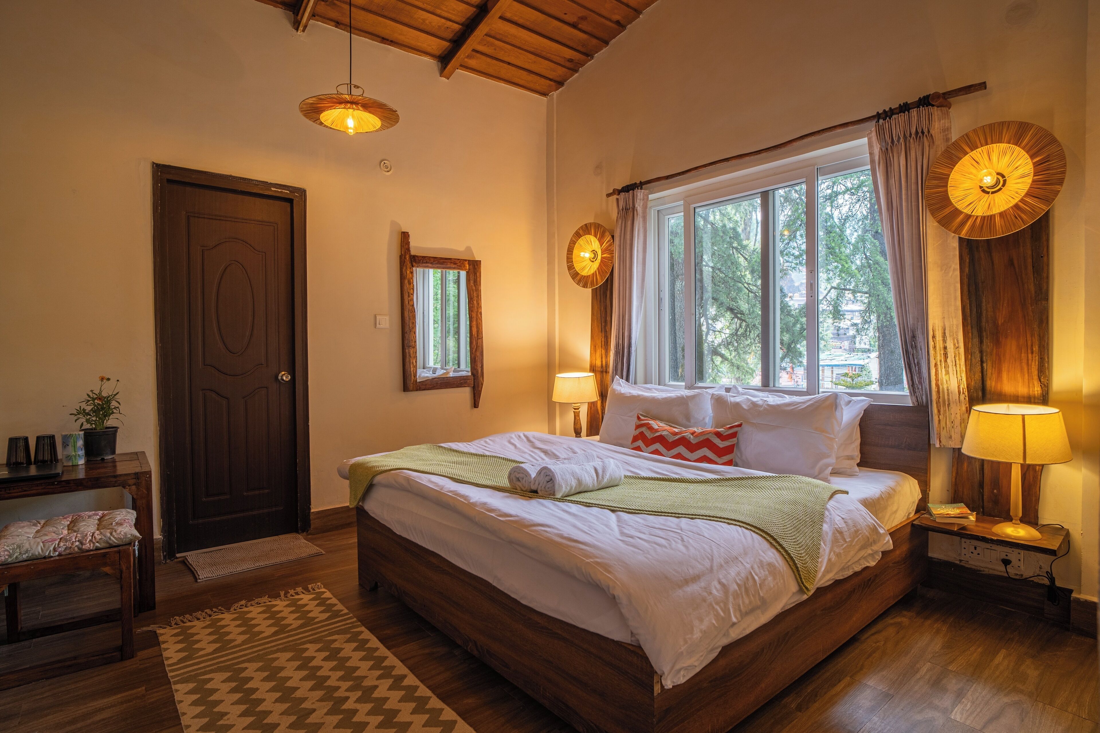 A bedroom with a wooden bed, bedside lamps, a window with curtains, a mirror, and a door.
