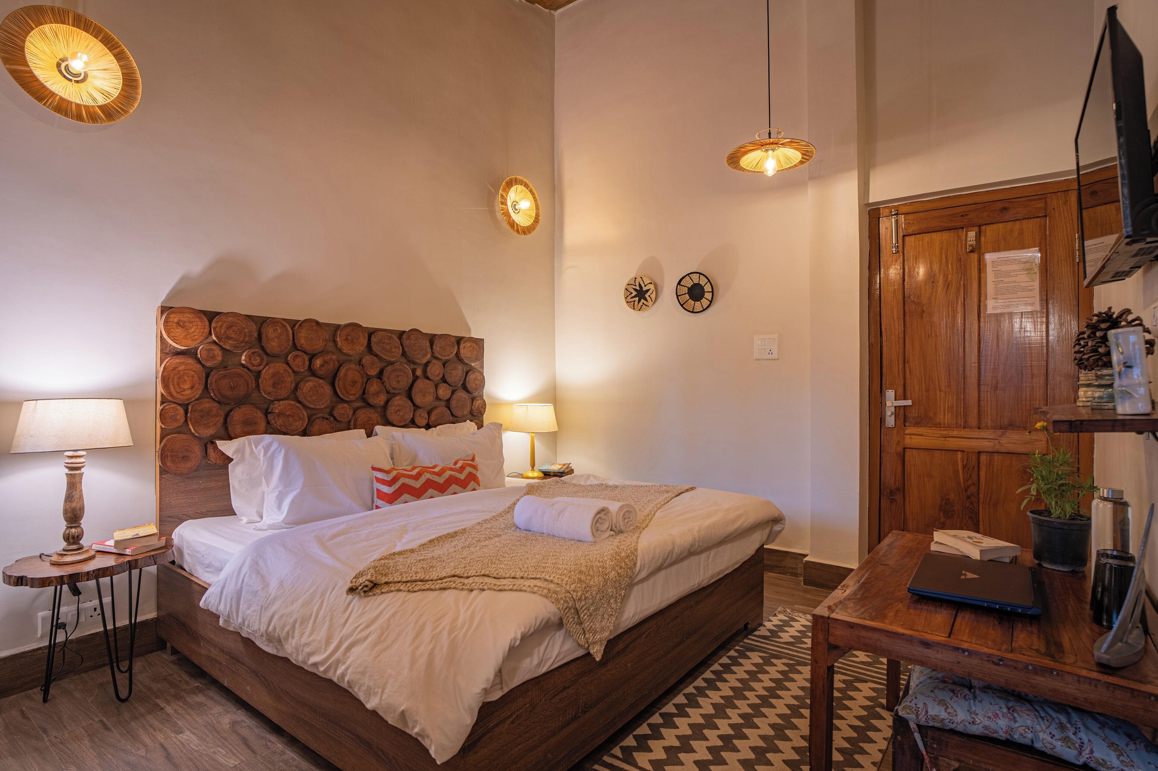 A bedroom with a wooden headboard, two bedside lamps, a bed with white bedding, a small table with a laptop, and a wooden door.