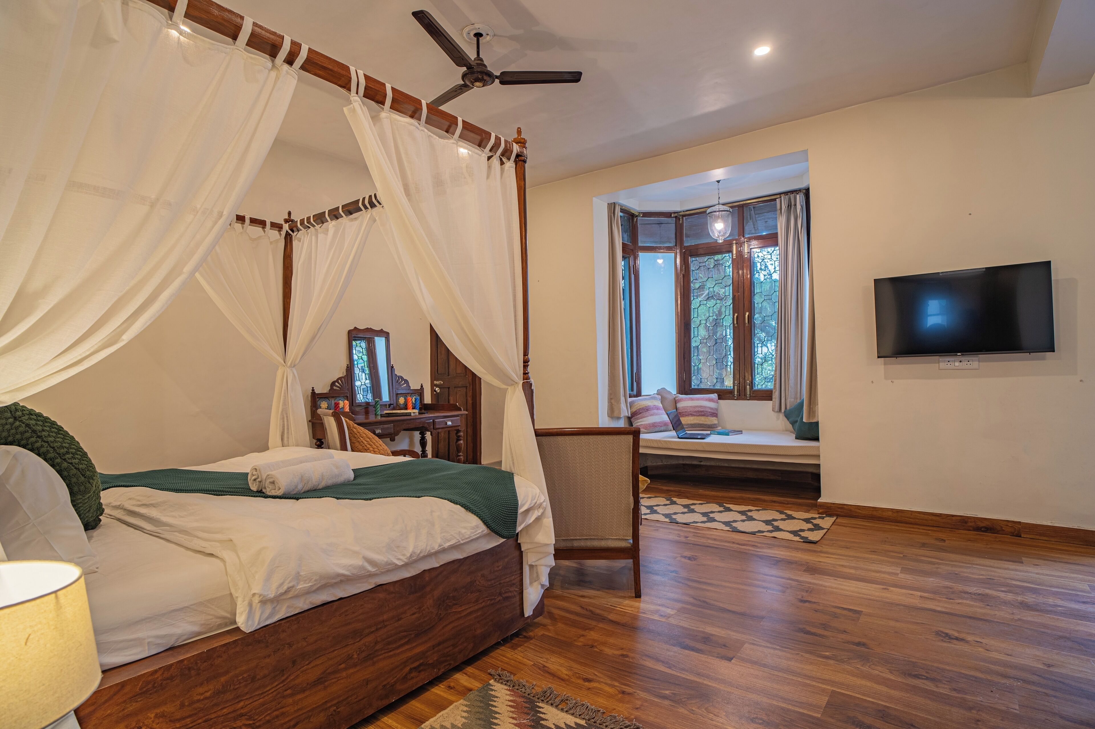A bedroom with a four-poster bed, a ceiling fan, a TV, and a window with a view of trees.