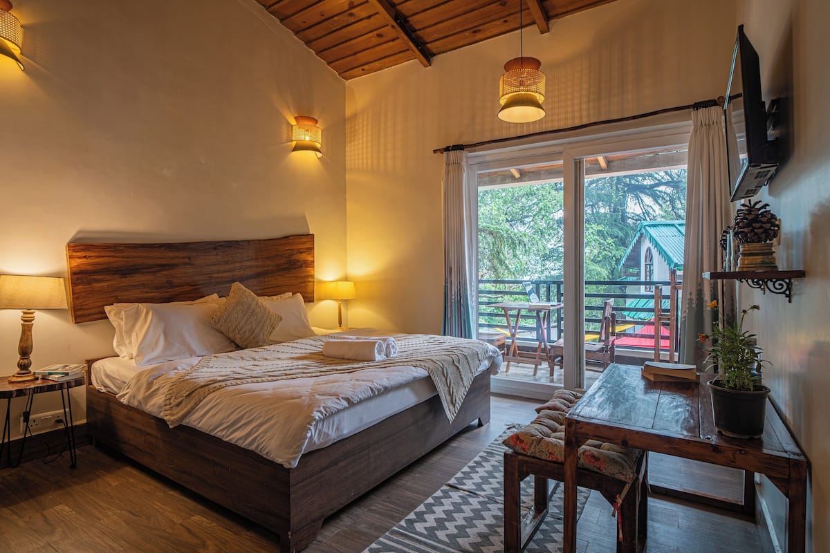 Une chambre avec une tête de lit en bois, un lit recouvert de draps blancs, une table de chevet avec une lampe, une table en bois ornée d’une plante, et une vue sur un balcon avec une chaise rouge.