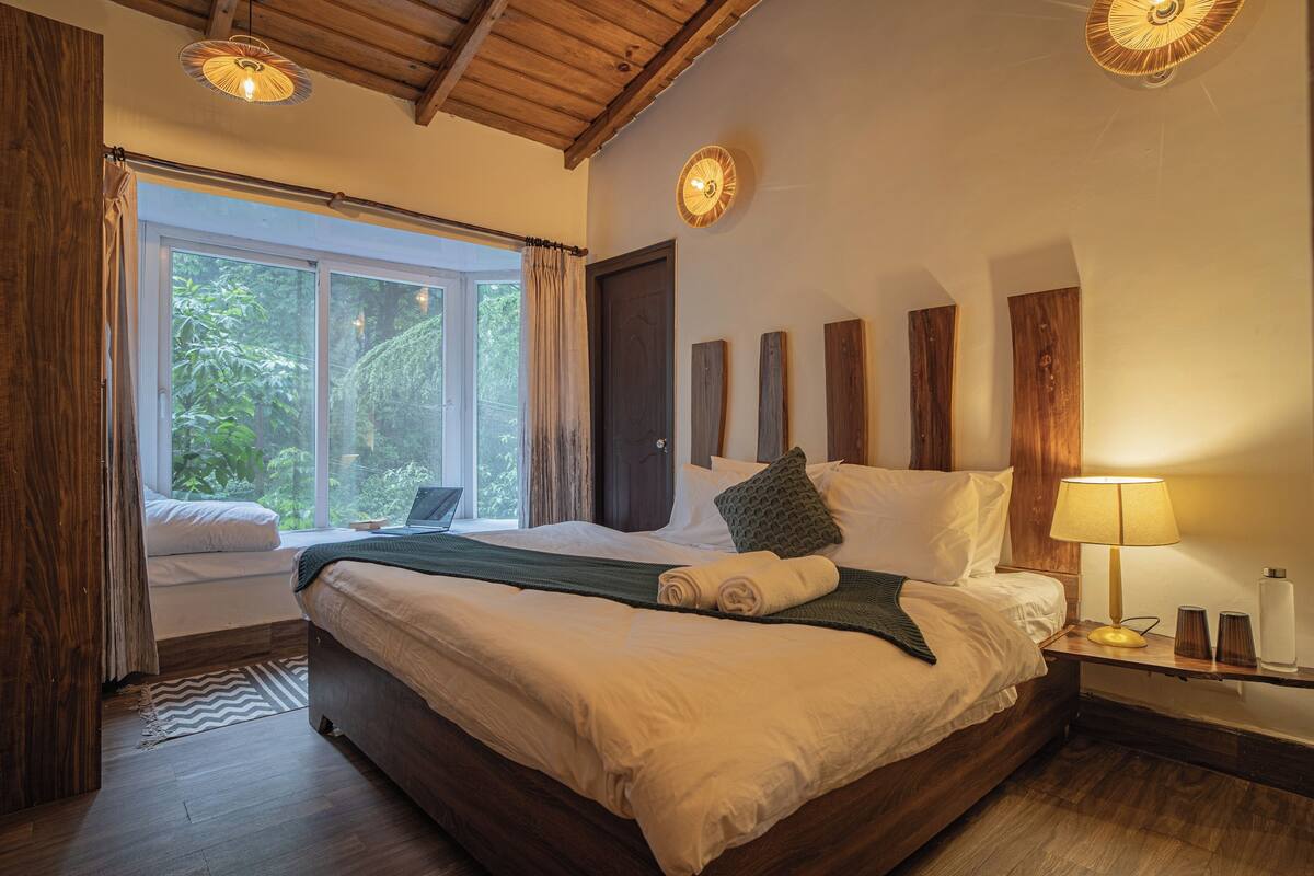 Une chambre avec un grand lit, une table de chevet avec une lampe, une fenêtre avec des rideaux et une vue sur la verdure à l’extérieur.