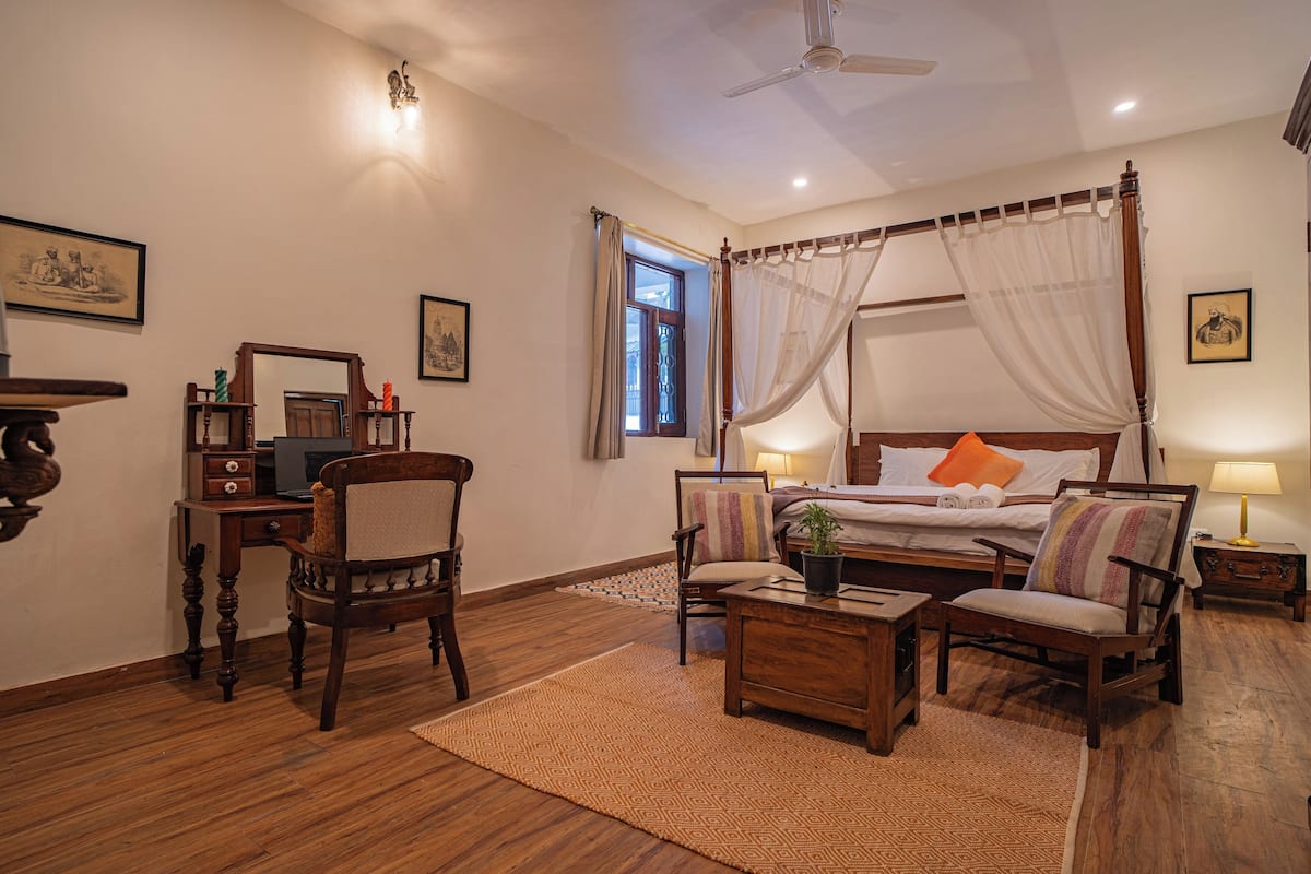 Une chambre à coucher avec un lit à baldaquin, un bureau, deux chaises, une petite table, une table de chevet, une lampe de chevet, un ventilateur de plafond et une fenêtre avec des rideaux.