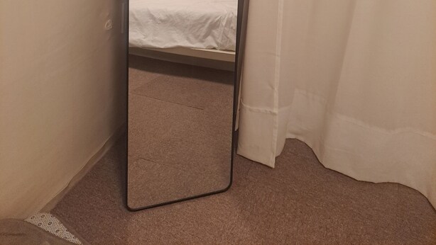 Un grand miroir étroit, doté d’un cadre noir, se trouve dans une pièce aux murs de couleur beige, où l’on aperçoit un lit recouvert de draps blancs.
