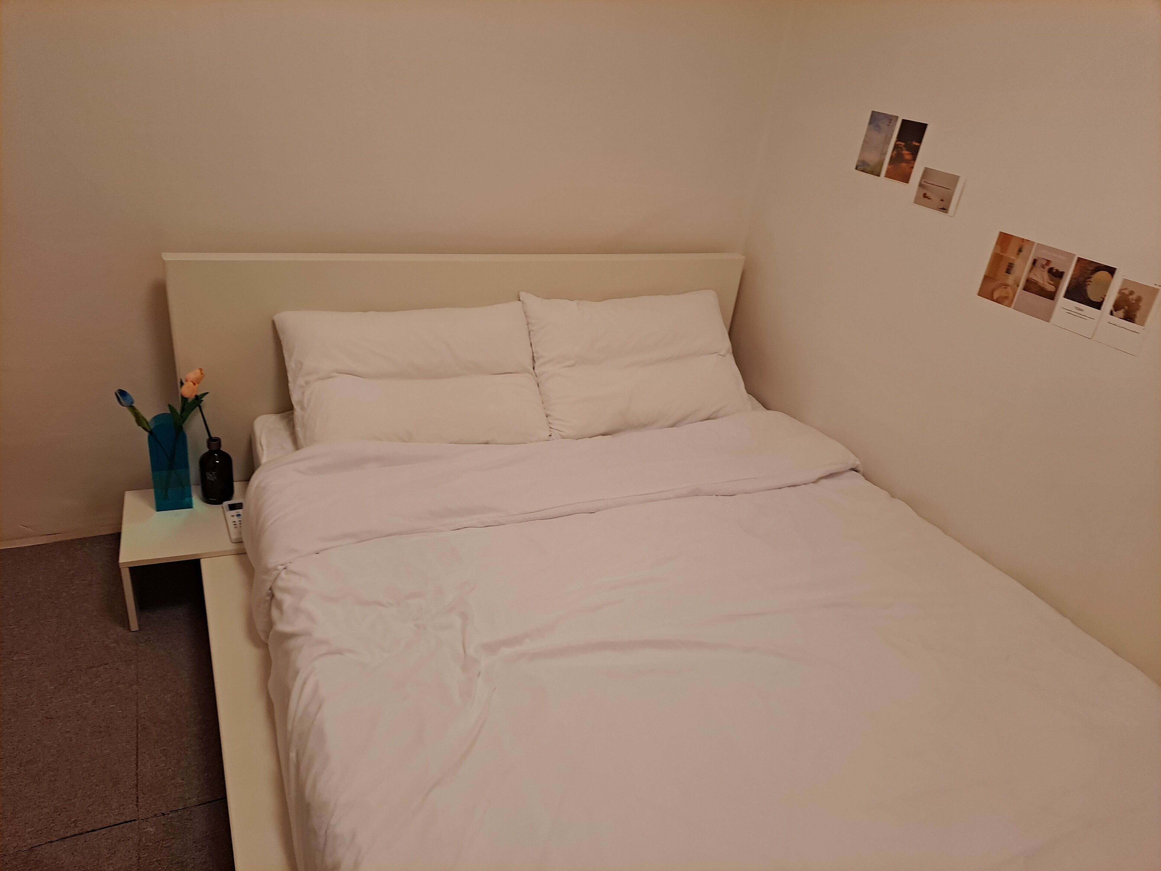 A neatly made bed with white bedding in a room with a plain wall and a small bedside table.