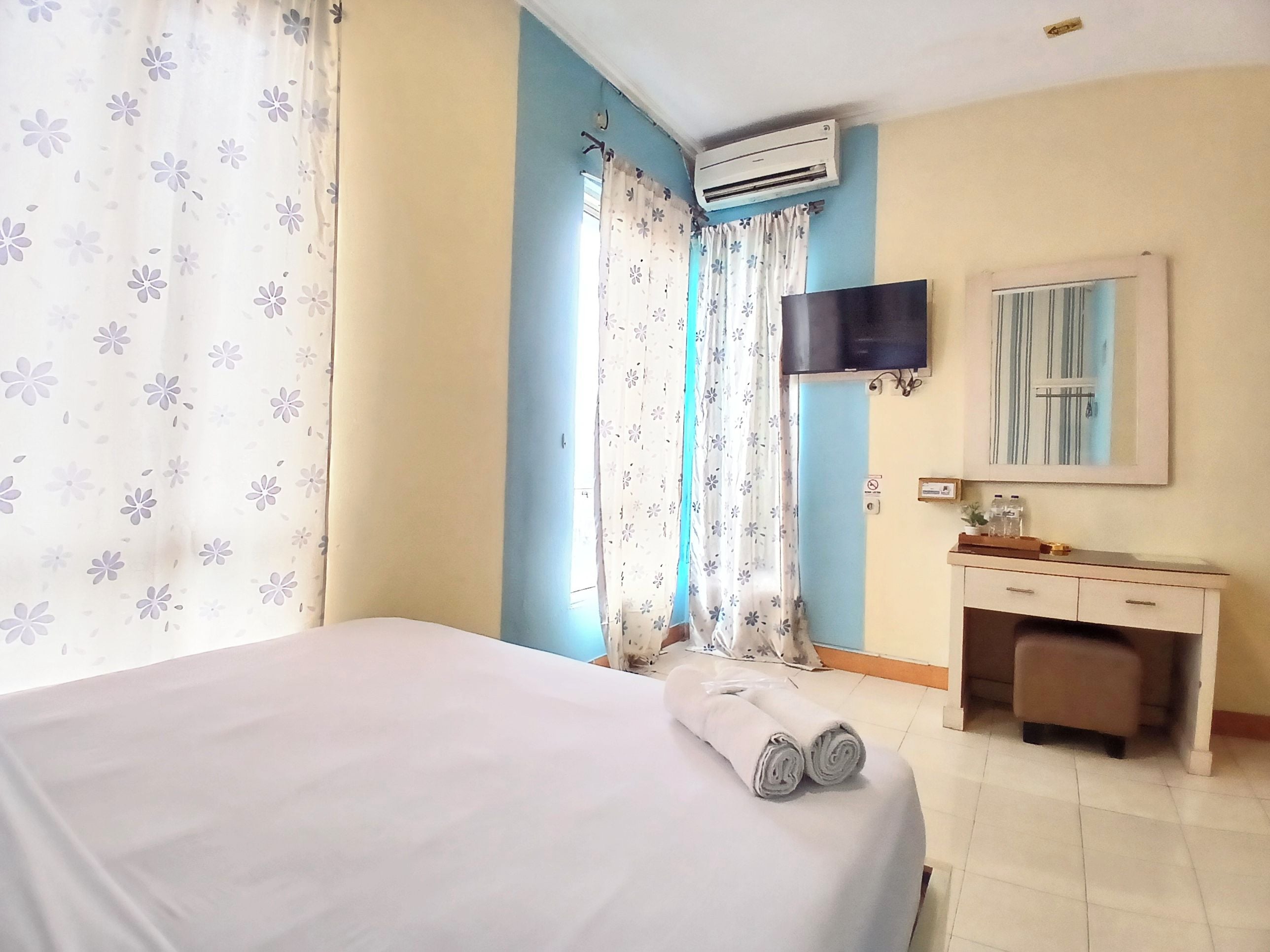 A hotel room with a bed, a bedside table, a mirror, an air conditioning unit, and curtains.