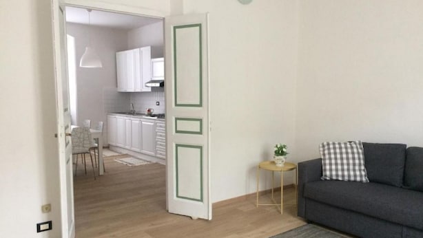 Una sala de estar moderna con un sofá gris y una mesita auxiliar con una planta. Se ve una cocina al fondo con armarios blancos y piso de madera.