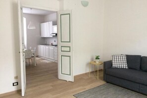 Una sala de estar moderna con un sofá gris y una mesita auxiliar con una planta. Se ve una cocina al fondo con armarios blancos y piso de madera.