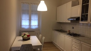 Una cocina compacta con armarios blancos, una encimera blanca, una mesa de comedor con sillas y una lámpara colgante.