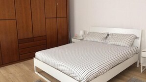 Un dormitorio con cama blanca, ropa de cama a rayas, un armario de madera y una mesita de noche con lámpara.
