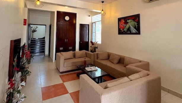 Uma sala de estar moderna com um sofá seccional, uma mesa de centro e plantas decorativas. Há um ventilador de teto, um ar-condicionado fixado na parede e um quadro emoldurado.