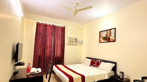 Quarto de hotel com cama, escrivaninha, cadeira, televisão, aparelho de ar condicionado e ventoinha de teto.