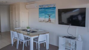 Een kamer met een eettafel, witte stoelen, een flatscreentelevisie en een schilderij van een pier.