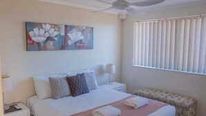 Een hotelkamer met een bed, nachtkastje, lamp, plafondventilator, raam met jaloezieën en een schilderij aan de muur.