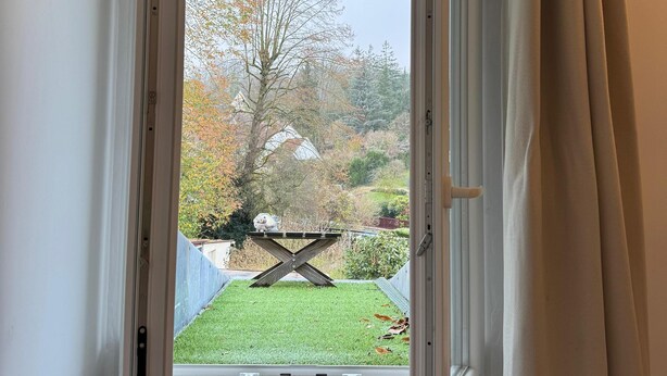 Une fenêtre avec un rideau entrouvert, d’où l’on aperçoit un espace verdoyant avec des arbres et une table en bois.
