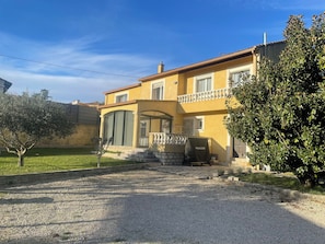 Une maison à deux étages avec une façade jaune, un escalier en pierre menant à l’entrée et un espace extérieur aménagé avec des sièges, protégé par un auvent.