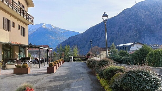 A café with outdoor seating, a mountain backdrop, and a street with a fence and greenery. - Edolo Apartment (Edolo)