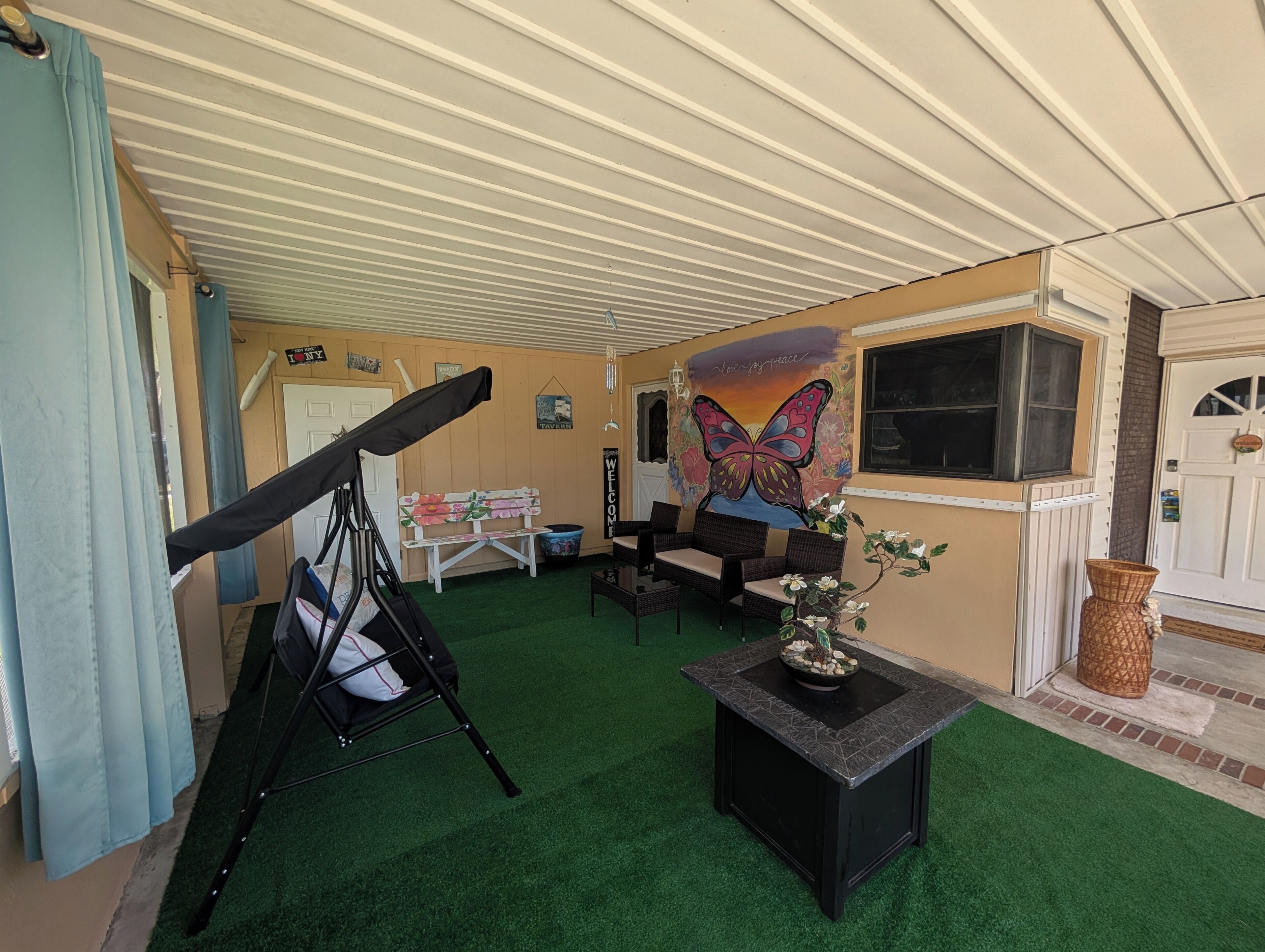 A room with a green artificial turf floor, a black folding chair, a white bench, wicker furniture, a wall mural, and a window with a view.