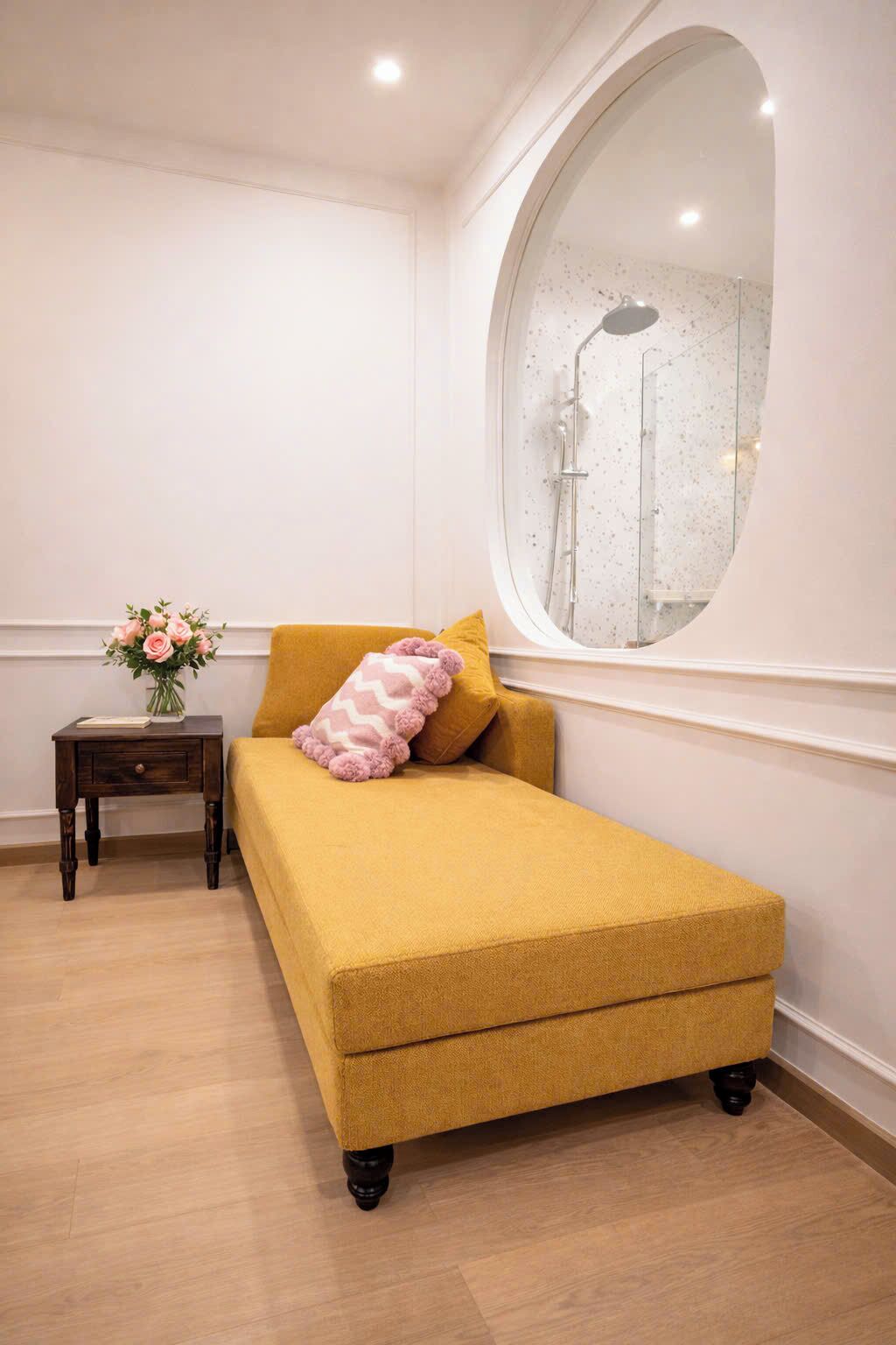 Un lit simple avec une couette de couleur moutarde, une petite table de chevet ornée d’un vase de fleurs et un grand miroir accroché au mur.