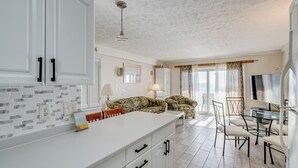 Amplia sala de estar con piso de baldosas blancas, una isla central con encimera blanca, un sofá y una mesa de comedor con sillas.