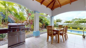 An outdoor patio with a grill, wooden table, and chairs, surrounded by lush tropical plants and a view of a swimming pool. - Beautiful 3-bedroom house in the exclusive, gated community of Chontales. (Chontales)